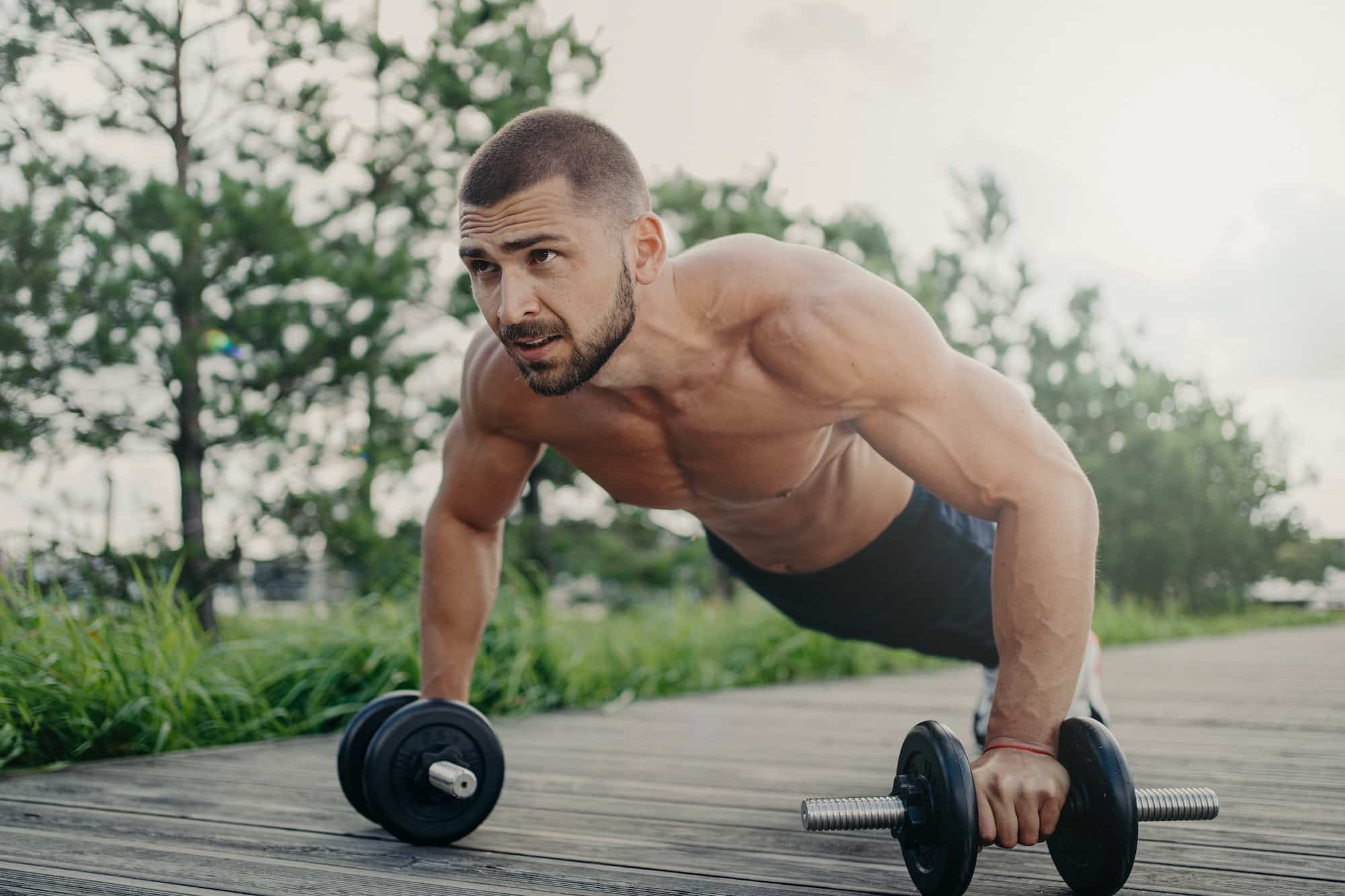 Muscular male bodybuilder does push up with barbells stands in plank pose with naked body.